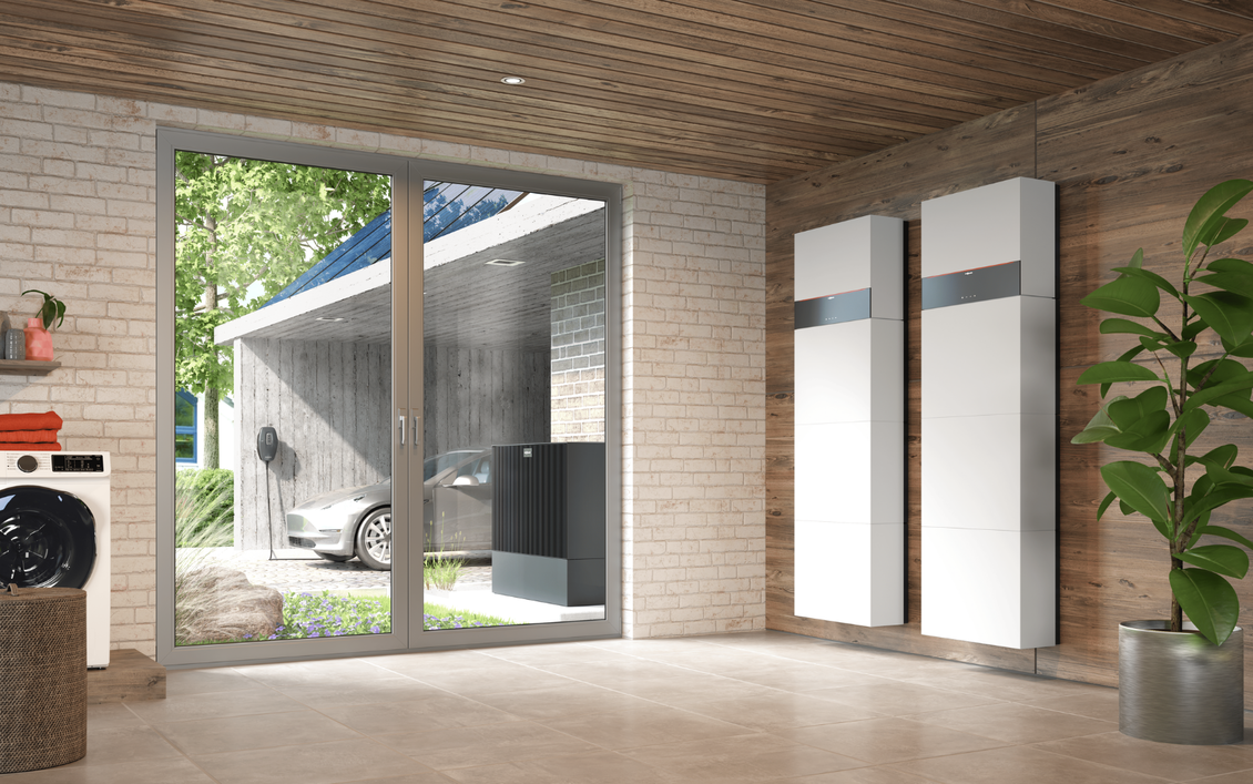 Hauswirtschaftsraum mit zwei wandmontierten Stromspeichern, Waschmaschine und Blick auf die Wärmepumpe und die Wallbox vor dem Haus.