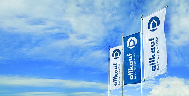 3 Fahnen mit dem Allkauf Haus-Logo vor einem blauen, leicht bewölkten Himmel